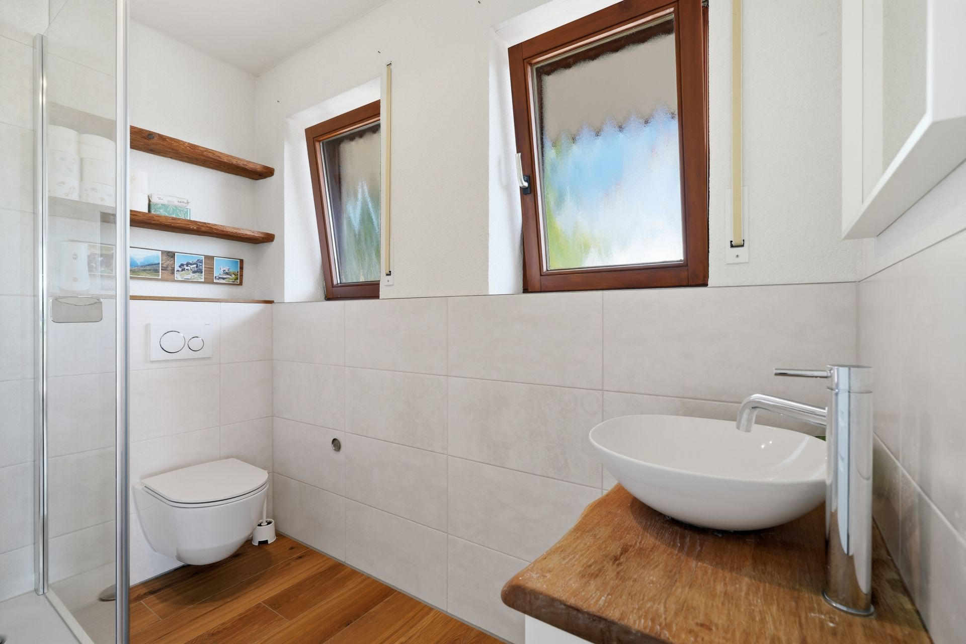 Modernes Badezimmer mit Wandtoilette, Waschbecken auf Holzplatte und zwei Fenstern.