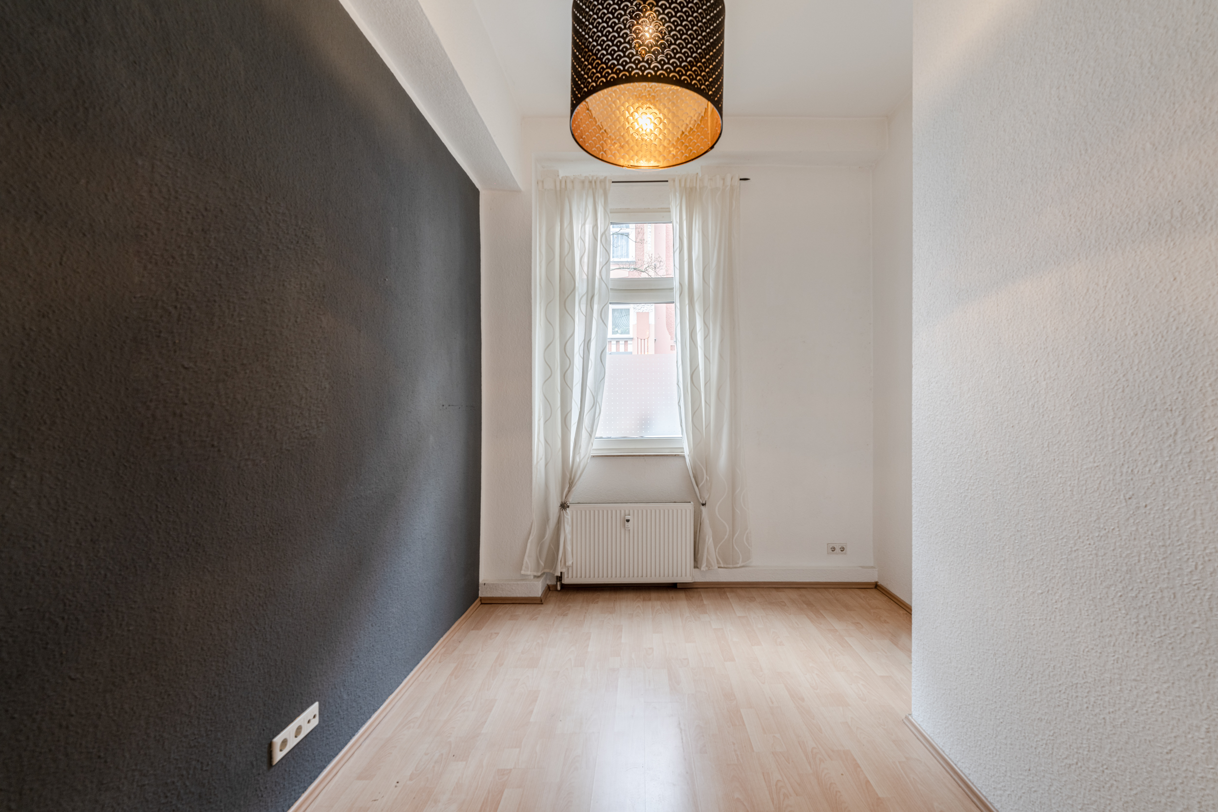 Leerer, schmaler Raum mit Holzfußboden, dunkler Wand, Fenster und weißem Vorhang.