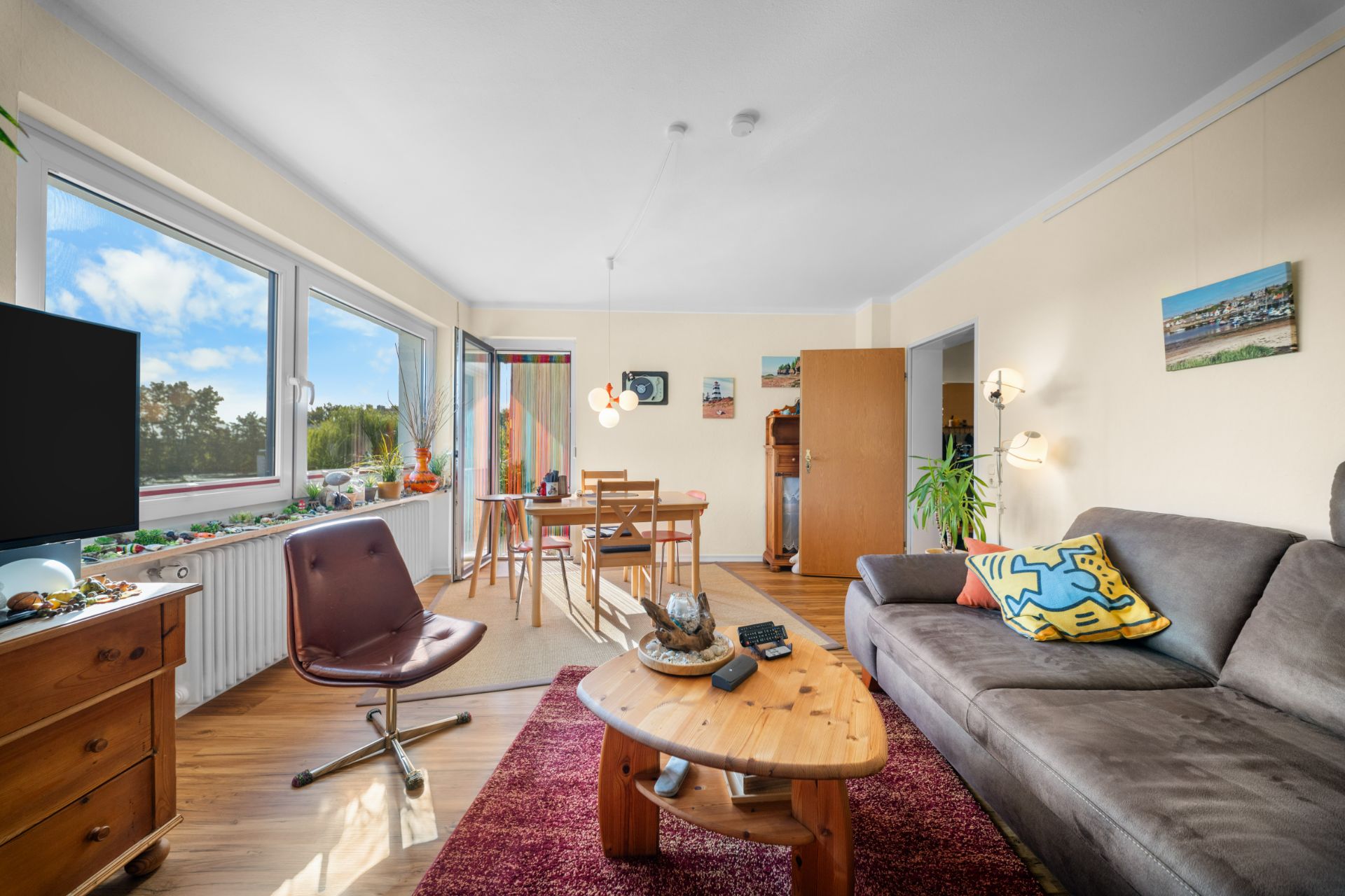 Helles Wohnzimmer mit Sofa, Couchtisch, Fernseher und Essbereich am Fenster.