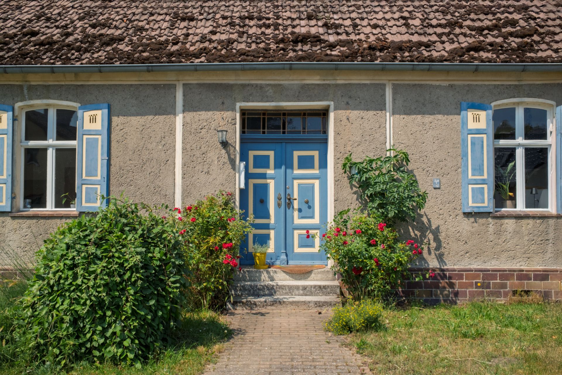 Eingang eines Hauses mit blauer Tür, Blumen und Sträuchern davor.