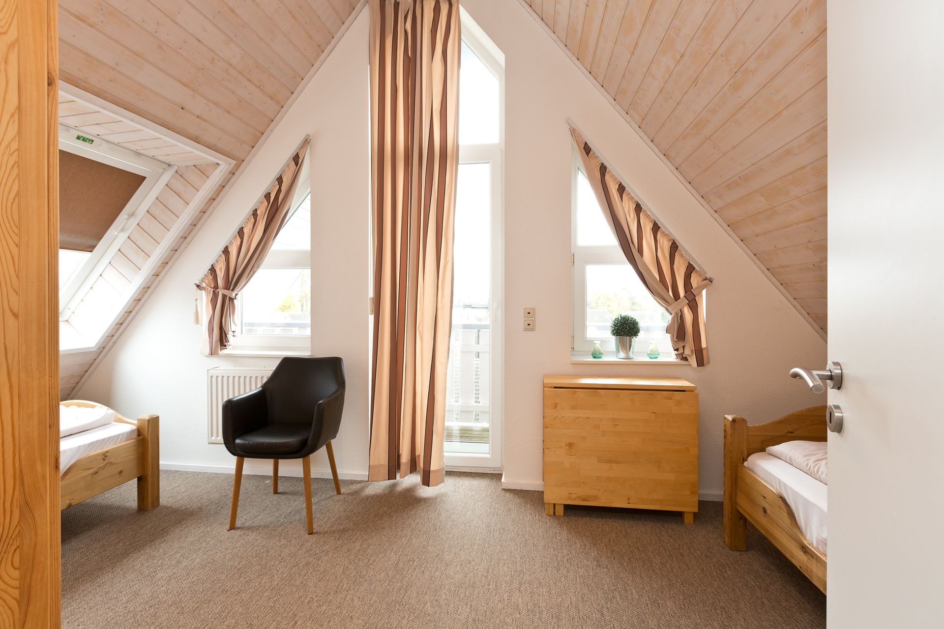 Helles Zimmer mit zwei Einzelbetten, einem Stuhl, Kommode und großen Fenstern mit Vorhängen.