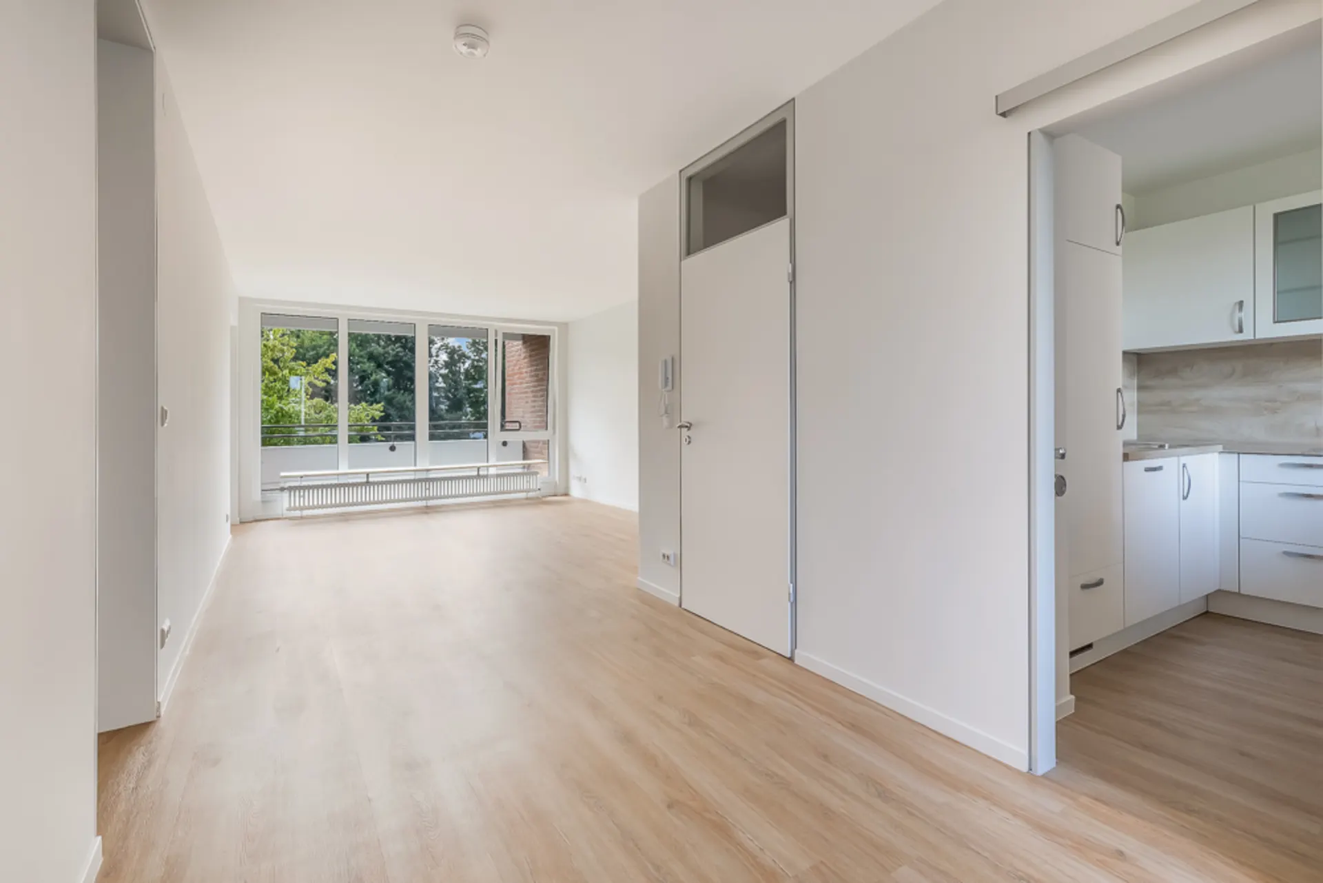 Helles, leeres Wohnzimmer mit großen Fenstern, Holzboden und Blick in eine weiße Küche.