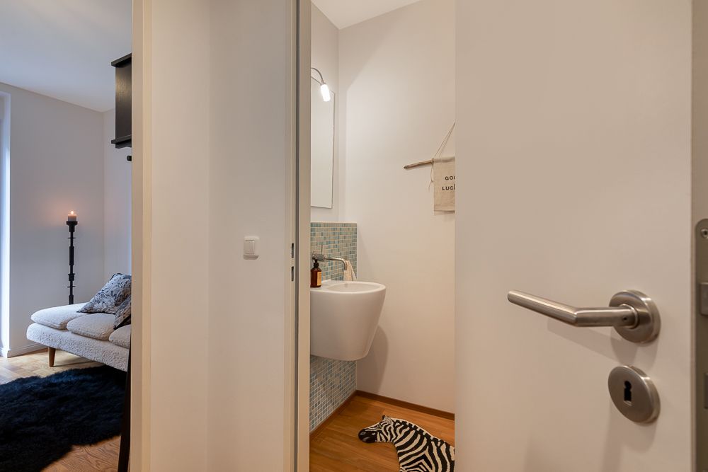 Offene Tür zeigt kleines Bad mit Waschbecken, daneben gemütliches Wohnzimmer mit Sofa und schwarzem Teppich.