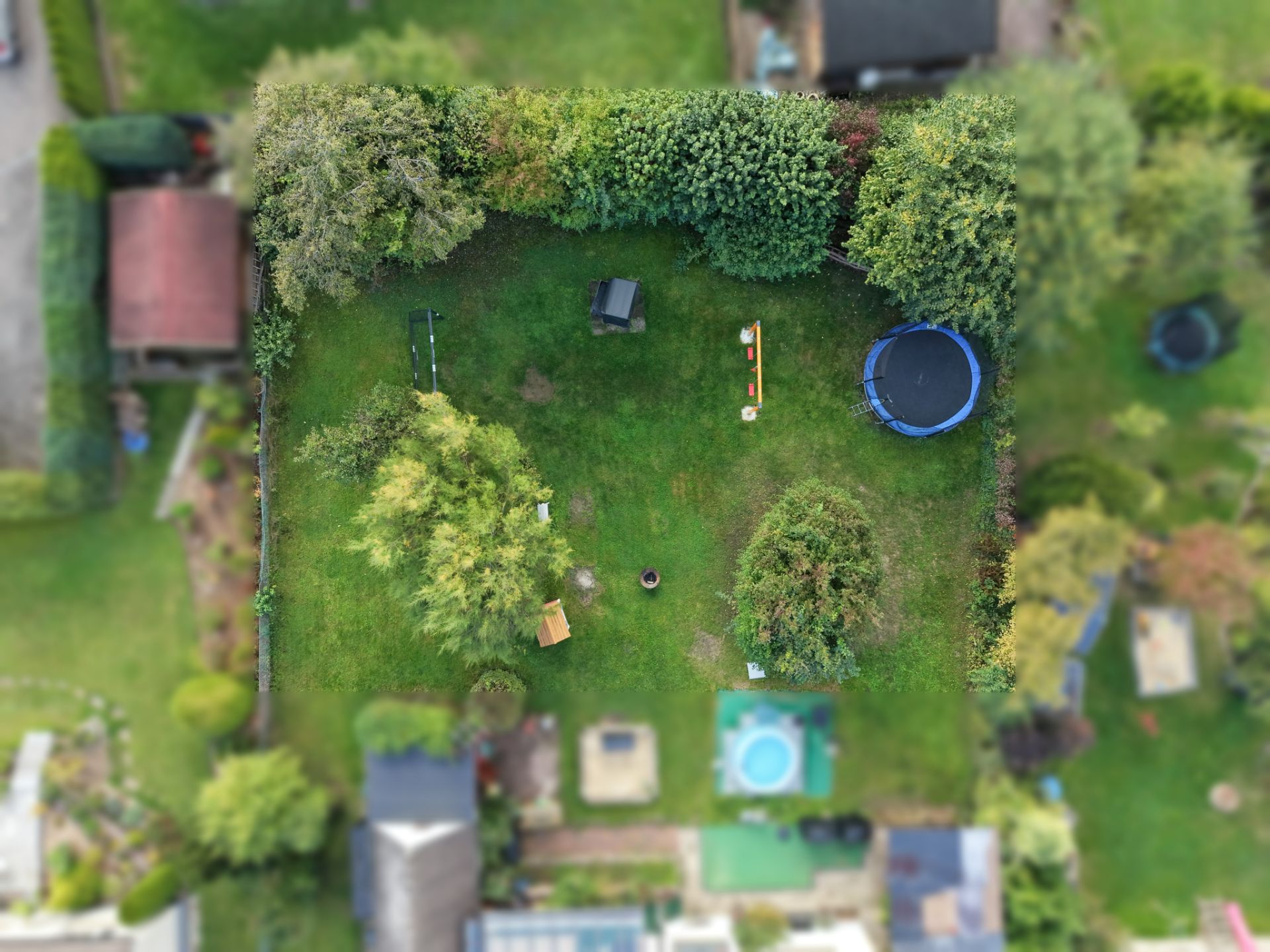 Luftaufnahme eines grünen Gartens mit Bäumen, Trampolin, Schaukeln und Sitzmöglichkeiten.