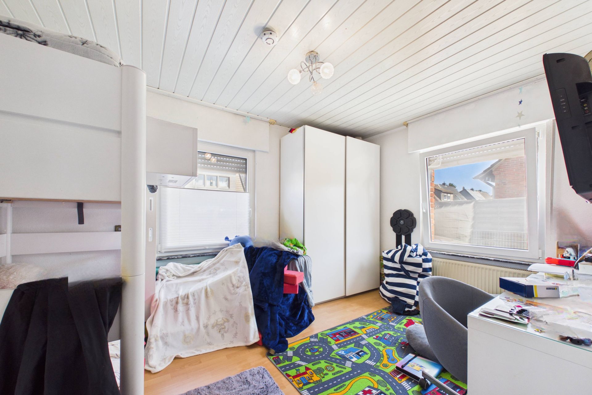 Helles Kinderzimmer mit Hochbett, Schrank, Schreibtisch und buntem Spielteppich.