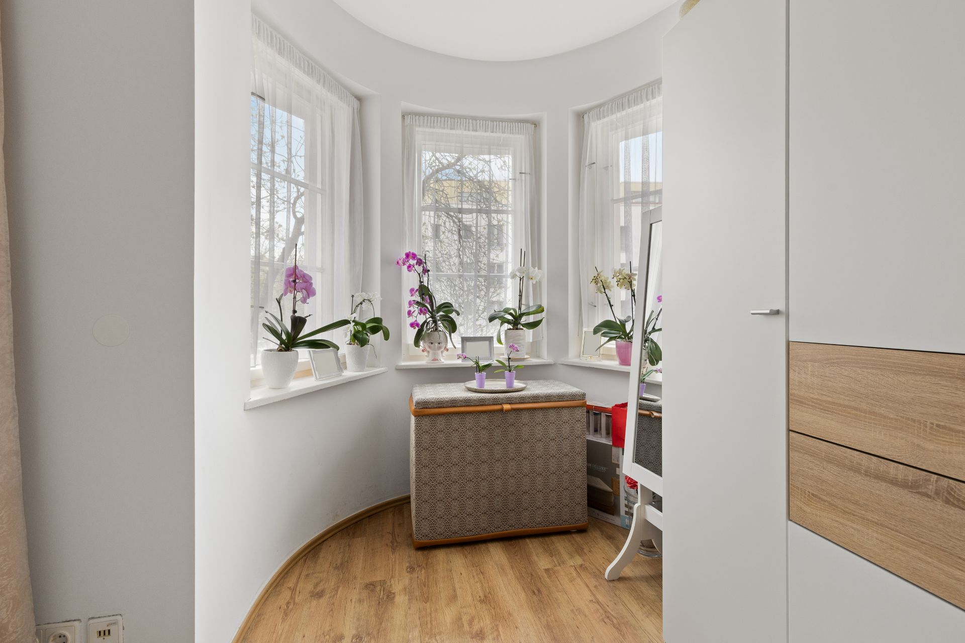 Heller Erker mit drei Fenstern, Blumen in Töpfen und einer Sitzbank auf Holzfußboden.