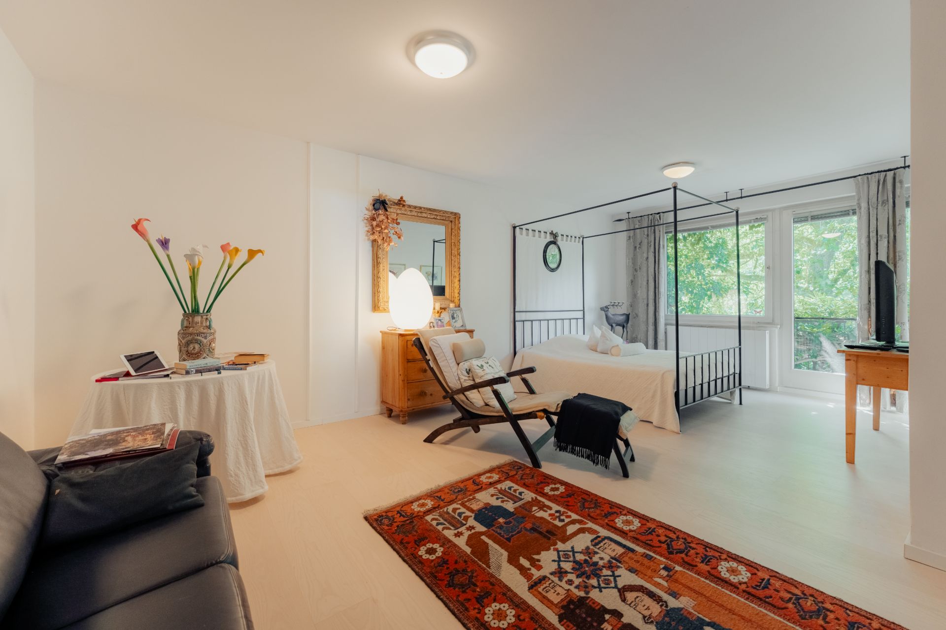 Helles Schlafzimmer mit Eisenbett, Teppich, Spiegel, Sessel und Schreibtisch vor Fenstern mit Blick ins Grüne.