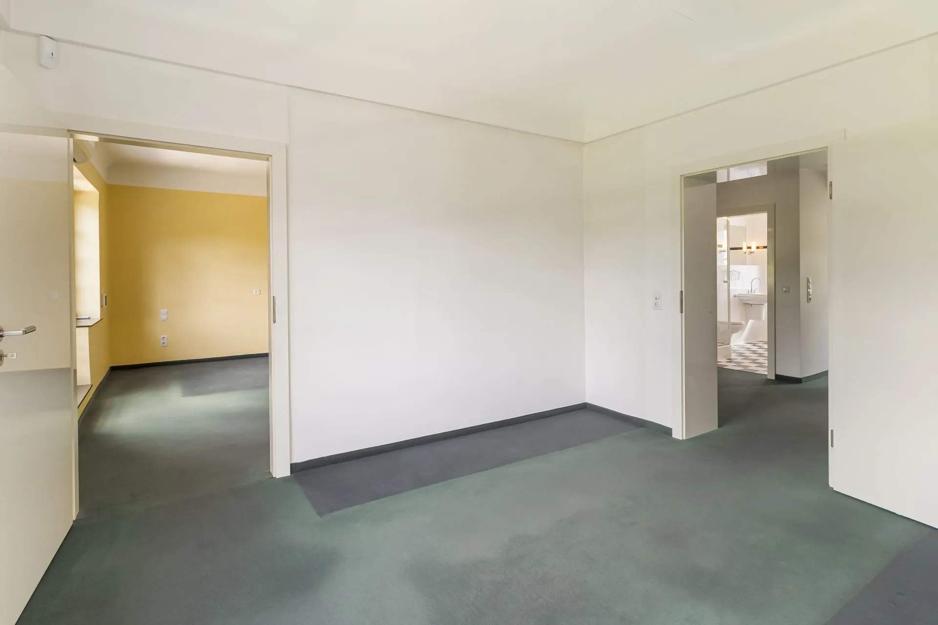Leerer Raum mit zwei offenen Türen, Sicht auf gelben Flur und Badezimmer mit Fliesenboden.