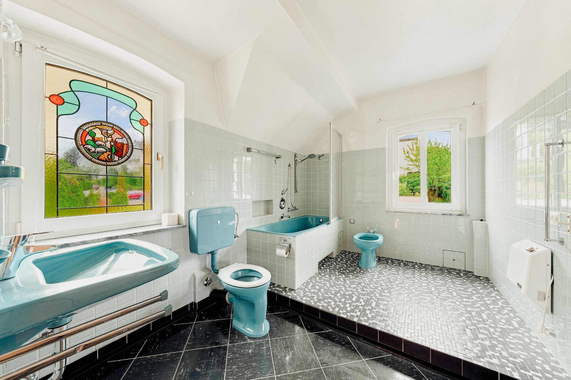 Helles Badezimmer mit blauen Sanitäranlagen, Badewanne, zwei Fenstern und Mosaikboden.
