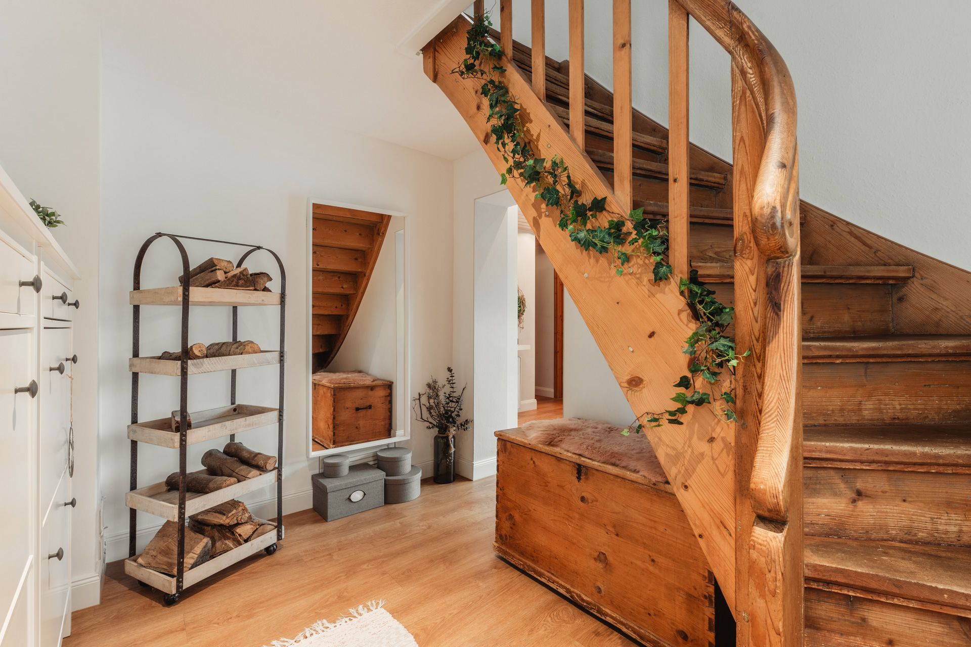 Holztreppe mit Efeudeko, Holztruhe und Regal mit Brennholz in einem Flur mit hellen Wänden.