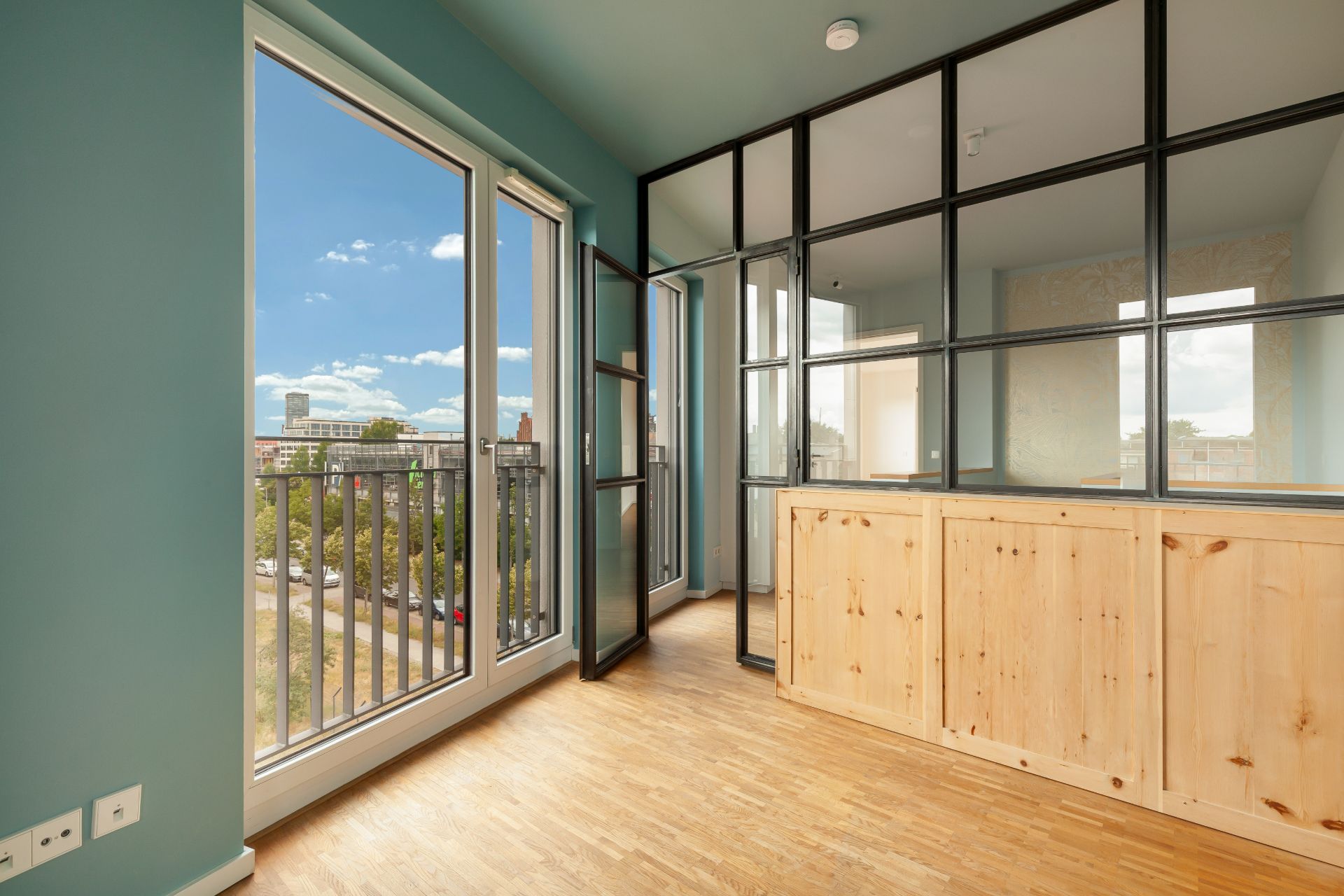 Helles Zimmer mit Holzboden, großer Fensterfront und Glaswand mit Holzverkleidung.