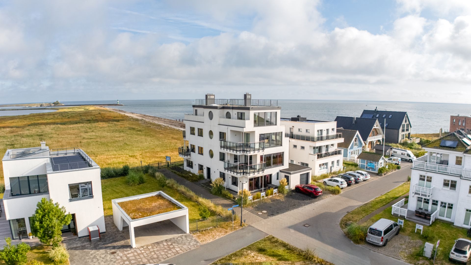 Moderne Wohnhäuser am Meer mit Balkonen und Autos auf der Straße, im Hintergrund Wiese und Wasser.