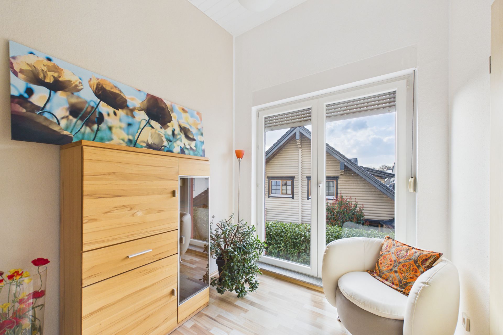 Helles Zimmer mit Schrank, Sessel, Pflanze und großem Fenster mit Blick auf ein Haus.