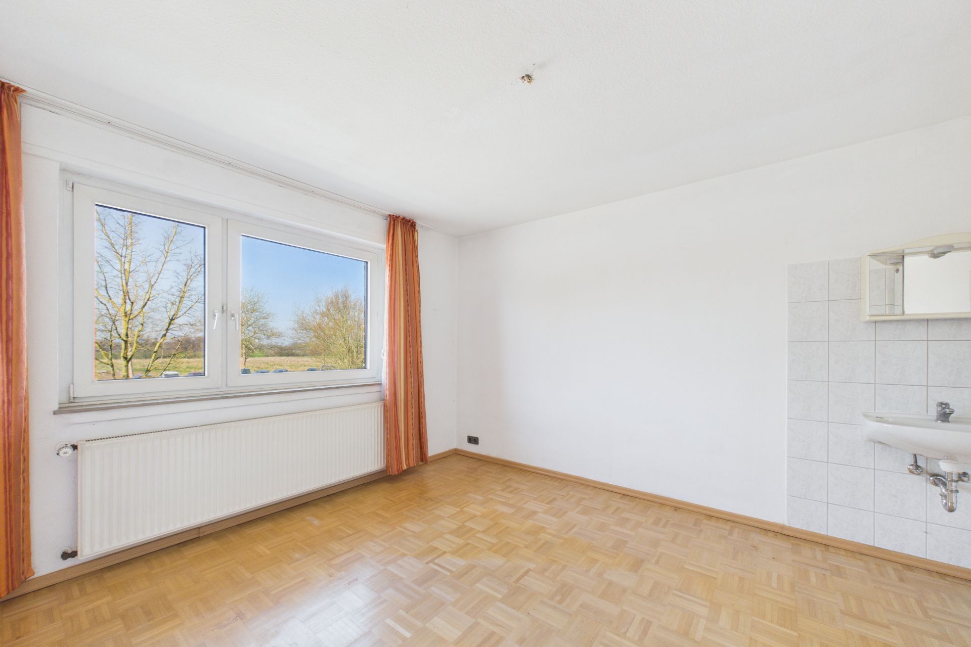 Helles Zimmer mit Parkettboden, großem Fenster, Waschbecken und Spiegel an der Wand.