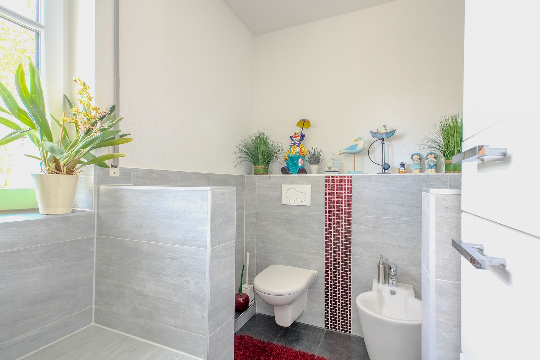 Modernes Badezimmer mit grauen Fliesen, WC, Bidet, Pflanzen und Dekofiguren auf der Ablage.