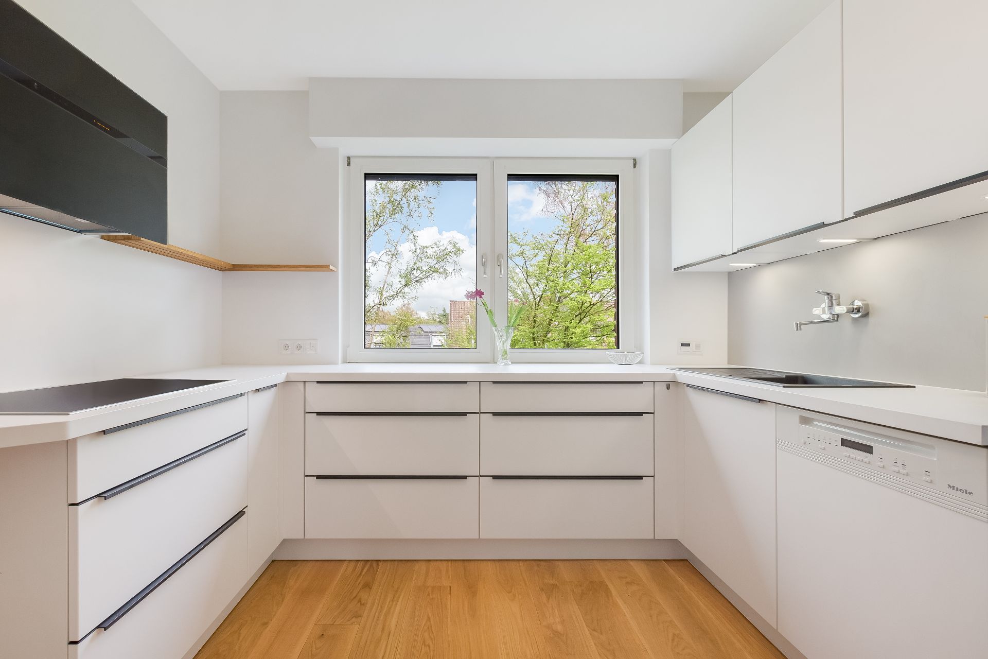Helle, moderne Einbauküche mit weißen Schränken, Holzfußboden und Fenster mit Blick ins Grüne.