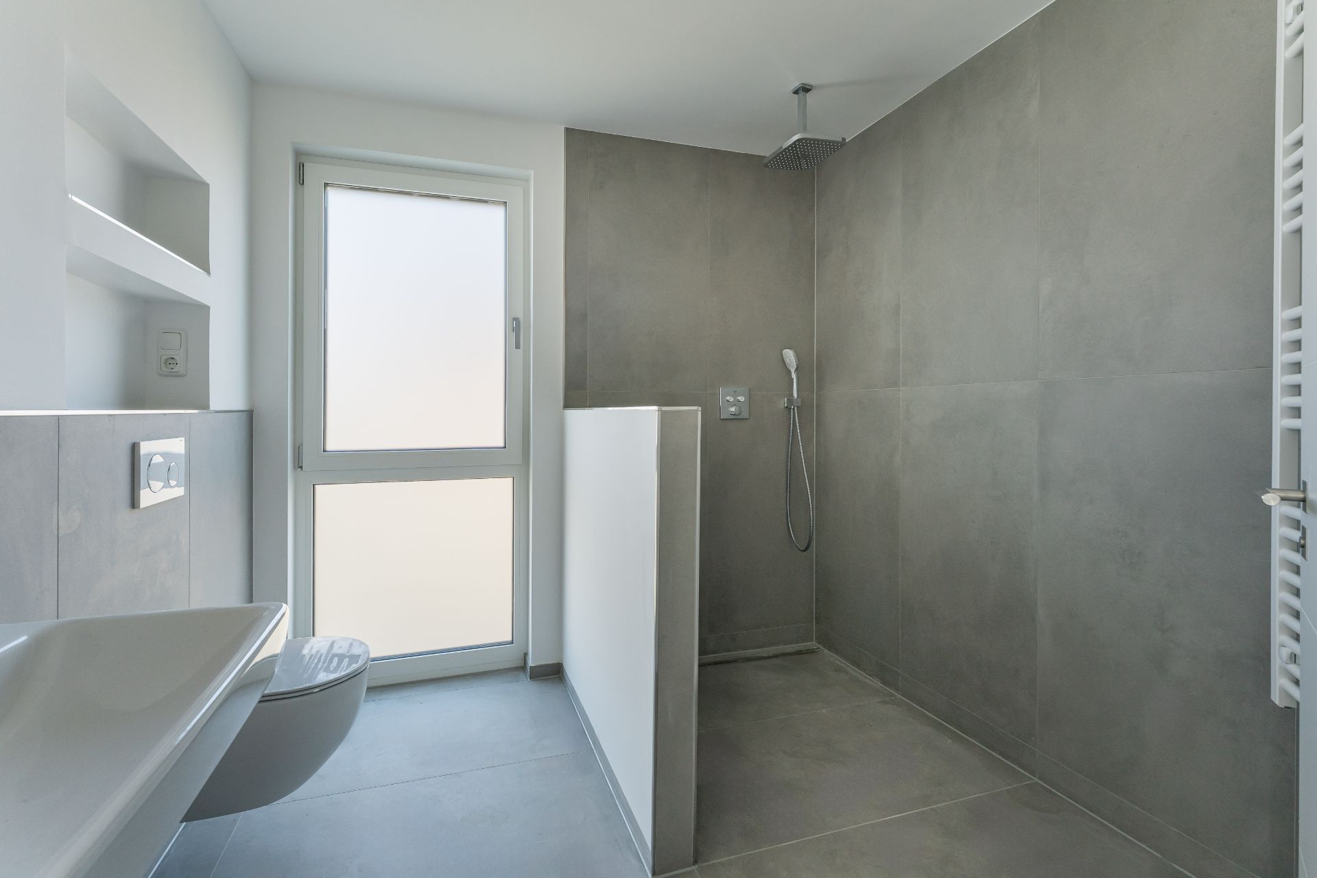 Modernes Bad mit bodengleicher Dusche, WC, Waschbecken und großem Fenster.