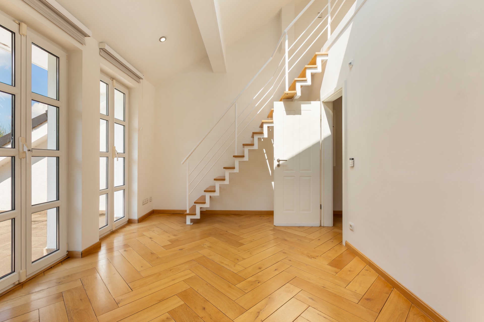 Helles Zimmer mit großem Fenster, Parkettboden und Treppe mit weißem Geländer.