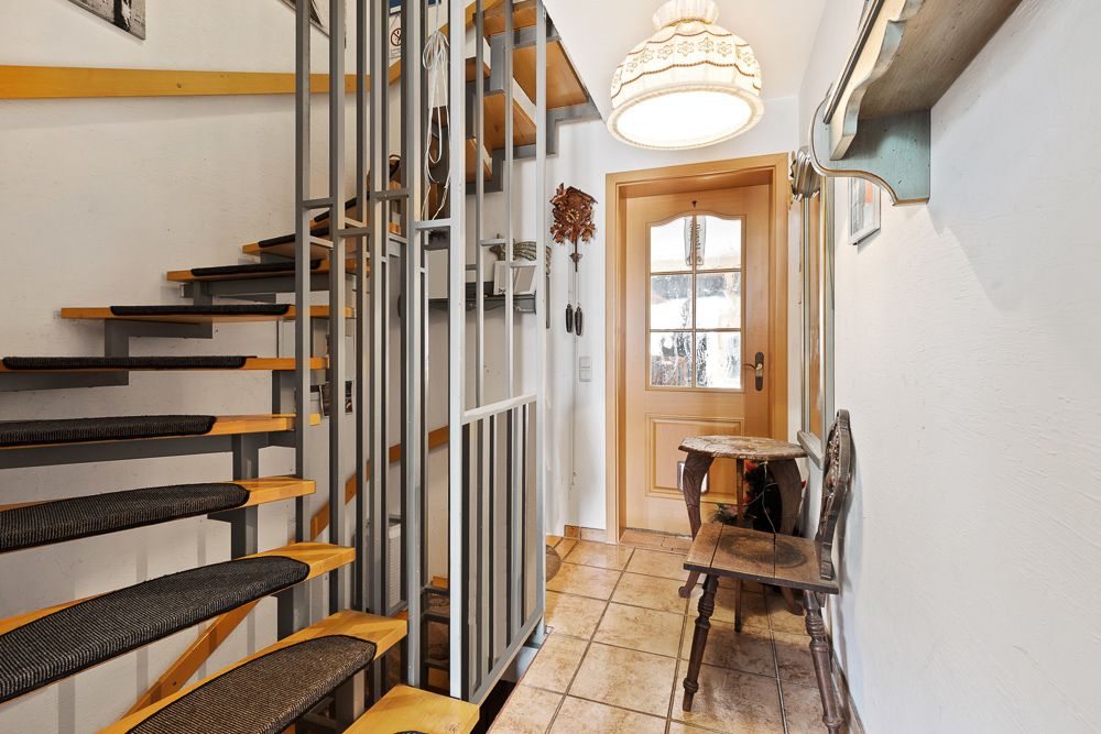 Flur mit Holztreppe, Eingangstür, Stuhl, kleinem Tisch und Deckenlampe.