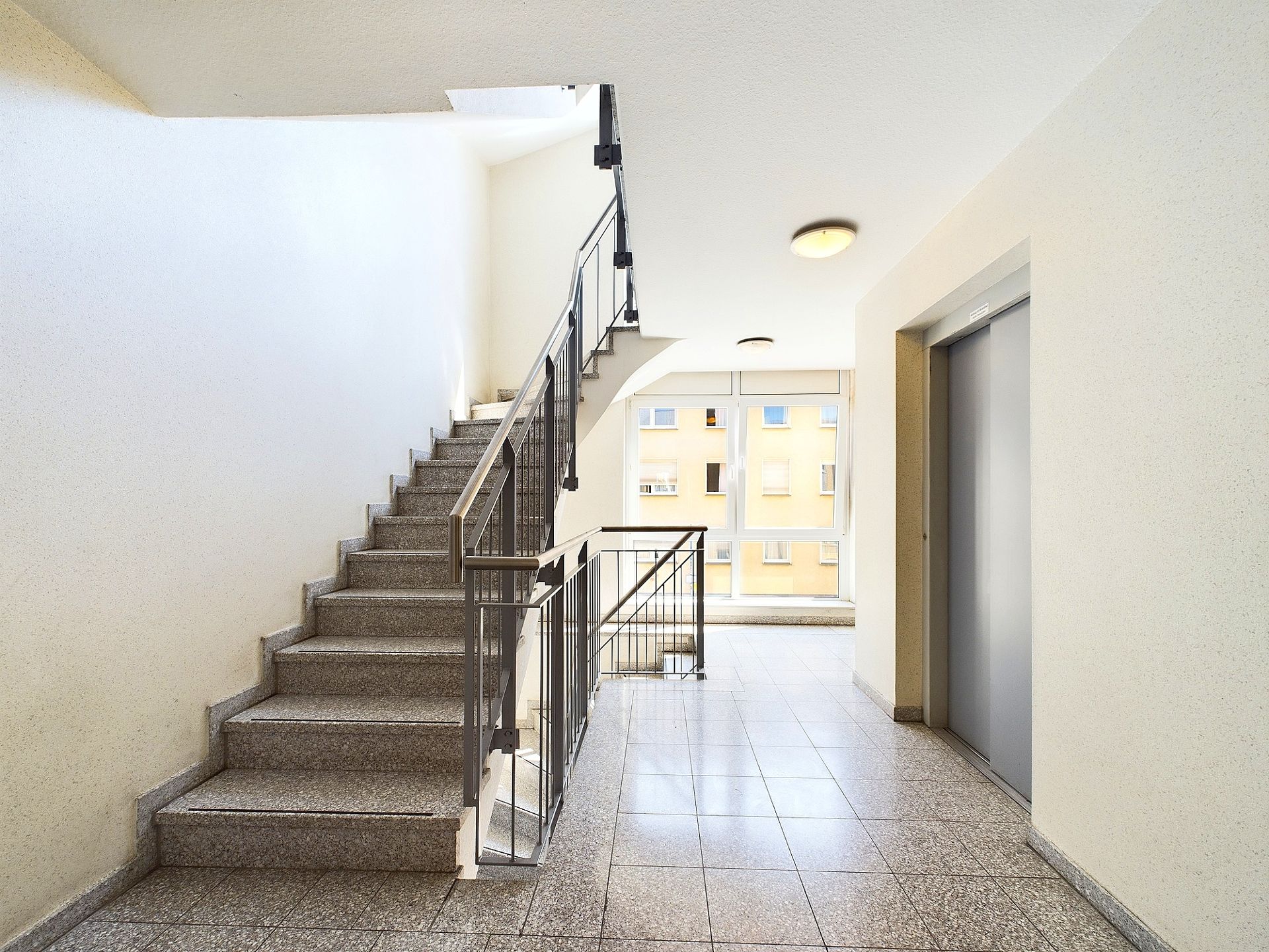 Treppenhaus mit Aufzug und großem Fenster in einem Wohngebäude.