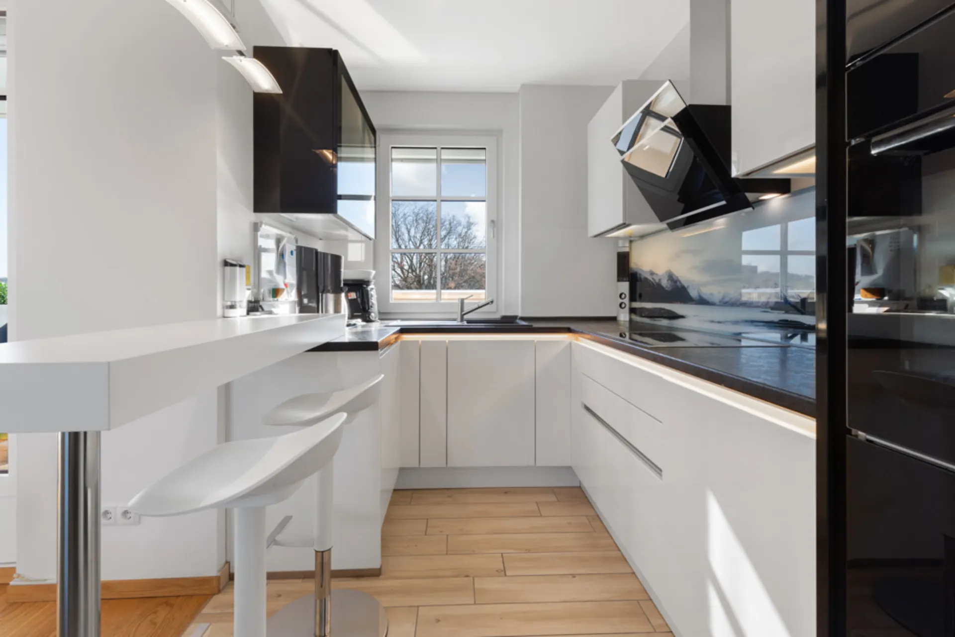 Moderne Küche mit weißen Schränken, Barhockern und großem Fenster im Hintergrund.