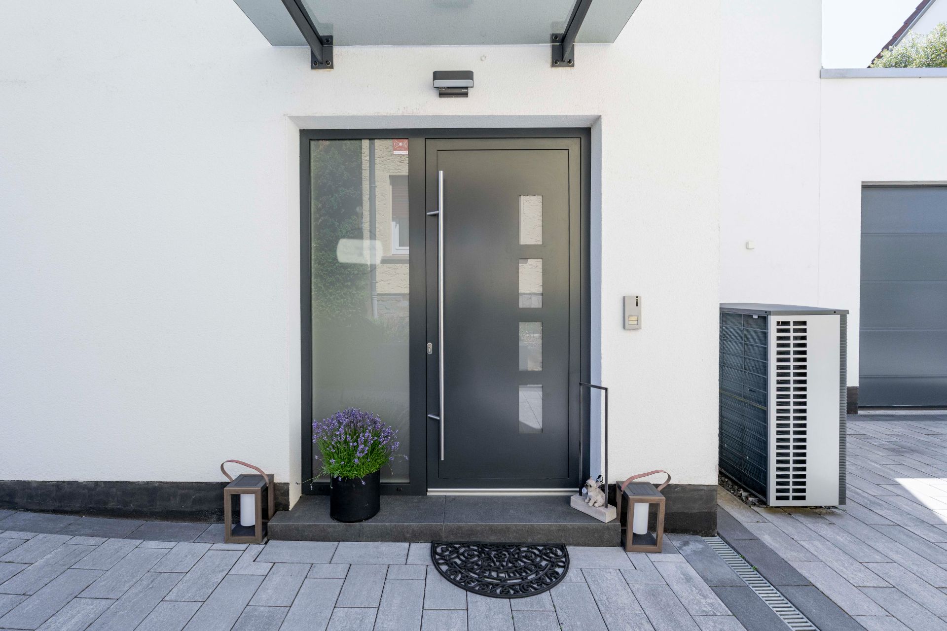 Moderne Haustür mit Glas-Seitenteil, Blumentopf und zwei Laternen daneben.