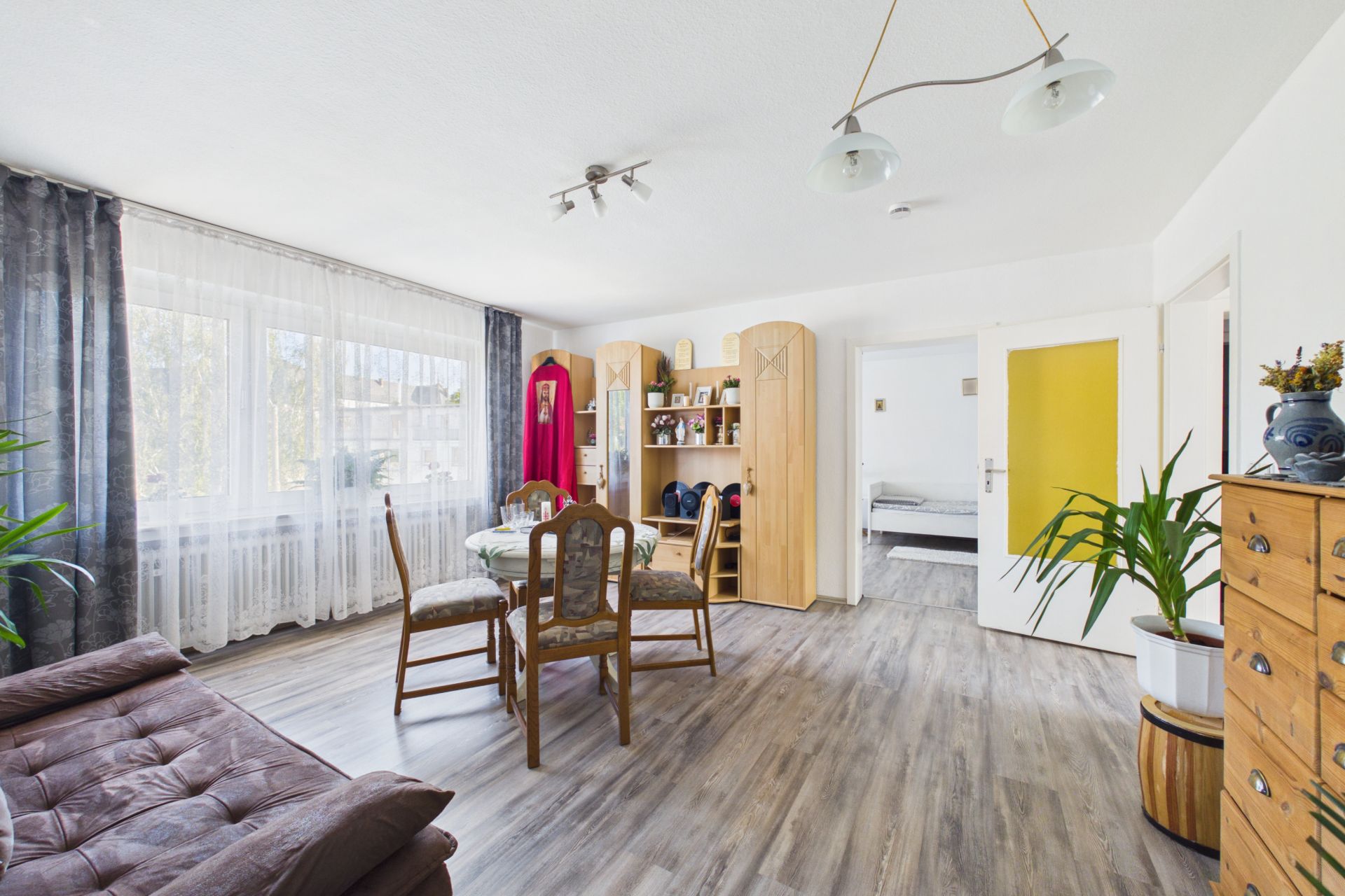 Helles Wohnzimmer mit Esstisch, Stühlen, Schrankwand, Sofa, Pflanzen und großen Fenstern.