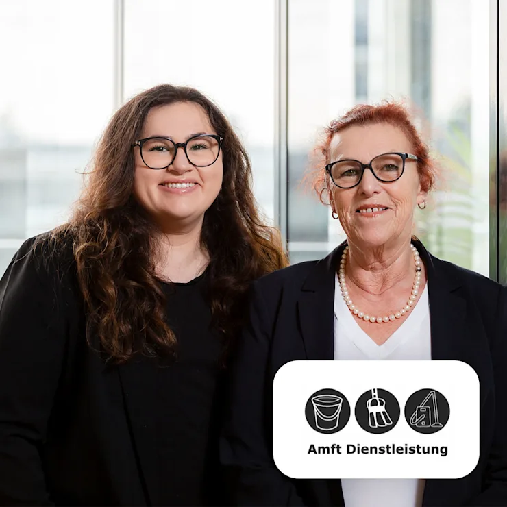 Zwei Frauen mit Brillen lächeln, stehen nebeneinander vor großen Fenstern.