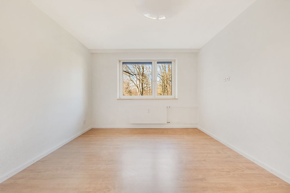 Leerer, heller Raum mit Holzfußboden und Fenster, durch das Bäume zu sehen sind.