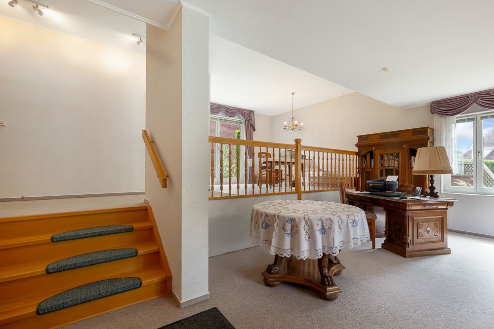 Wohnzimmer mit Holzmöbeln, rundem Tisch mit Decke, Schreibtisch, Fenster und Treppe.