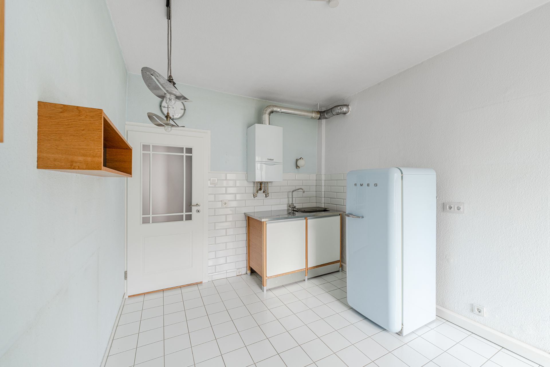 Helle, kleine Küche mit weißem Kühlschrank, Spüle, Hängeschrank und Fliesenboden.