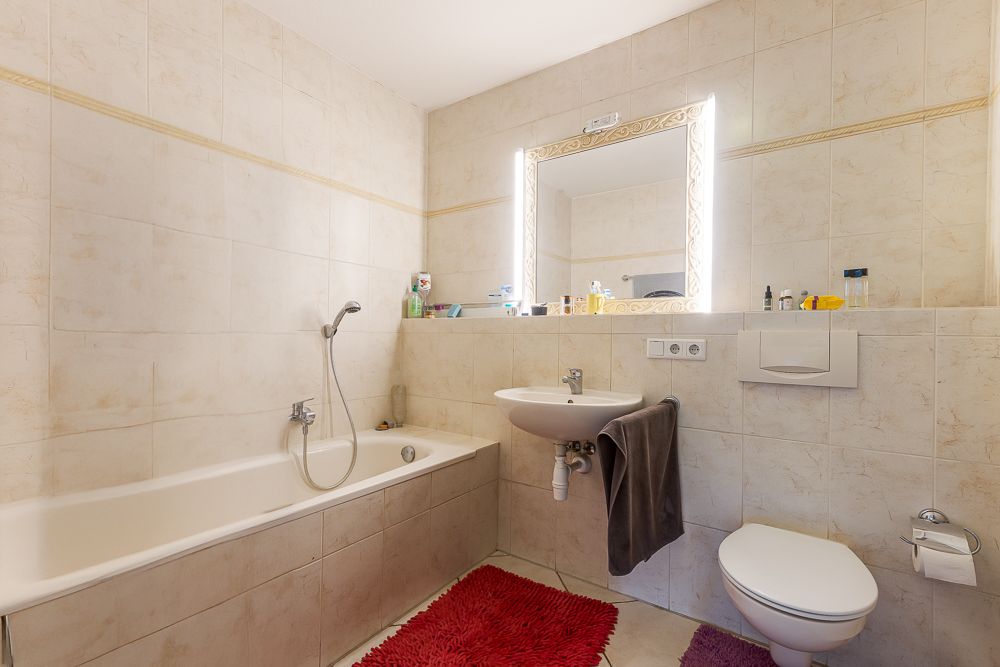 Helles Bad mit Badewanne, Waschbecken, Spiegel, WC und roten Teppichen auf hellen Fliesen.