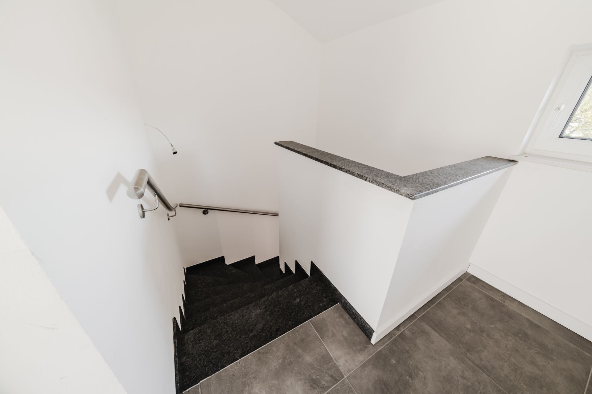 Schwarze Treppe mit Metallgeländer in einem weißen Flur, Fenster rechts neben dem Treppenaufgang.