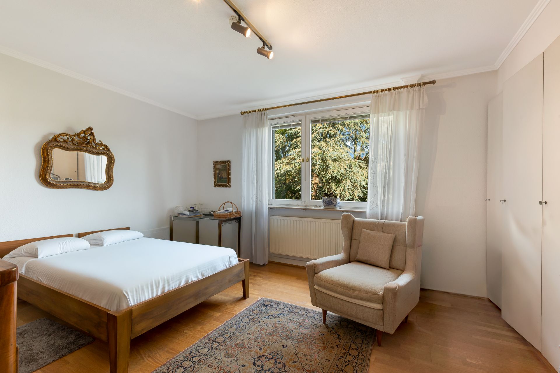 Schlafzimmer mit Doppelbett, Sessel, Fenster mit Vorhang und Holzfußboden.