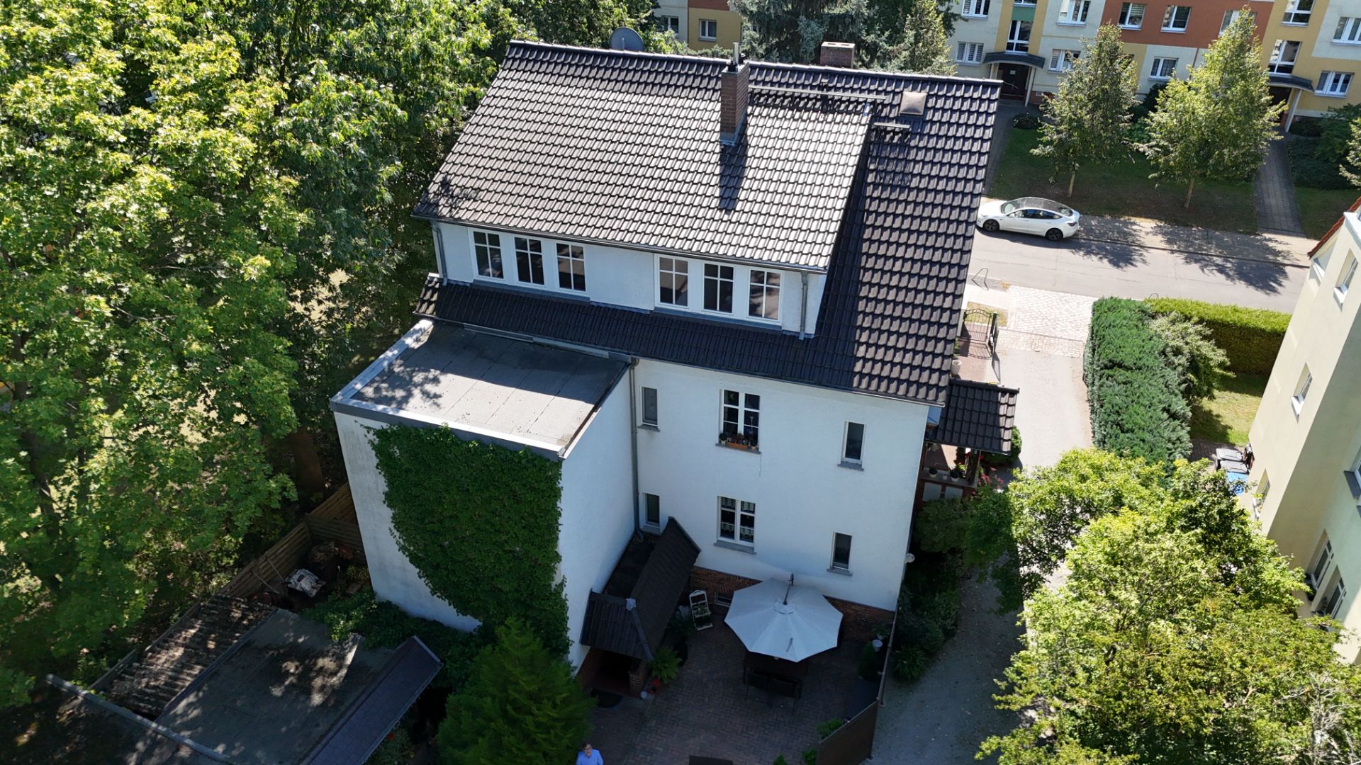 Luftaufnahme eines weißen Wohnhauses mit dunklem Dach und Garten mit Sonnenschirm.