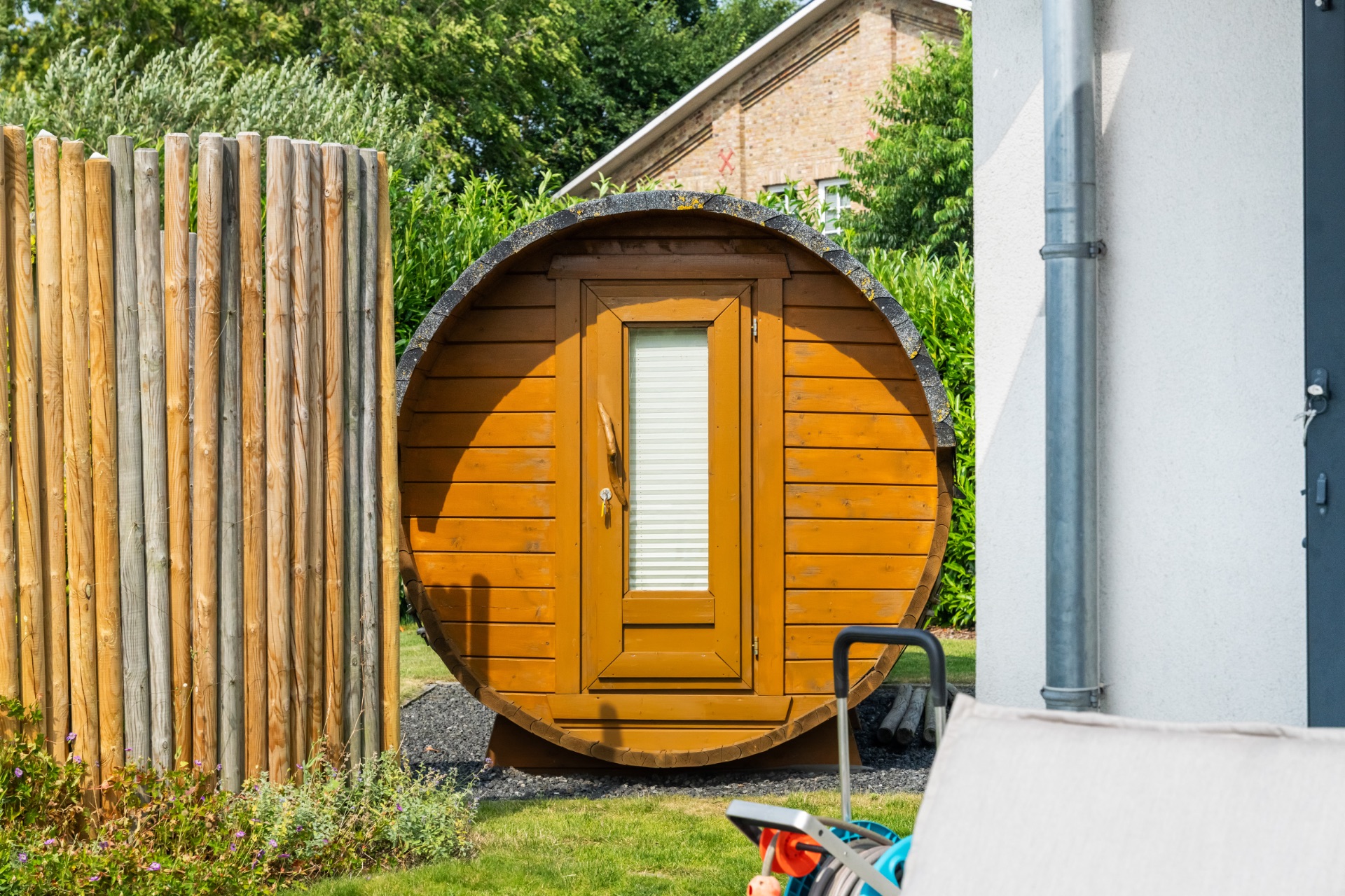 Eine runde, hölzerne Gartensauna mit Tür steht im Garten neben einem Sichtschutz aus Holz.