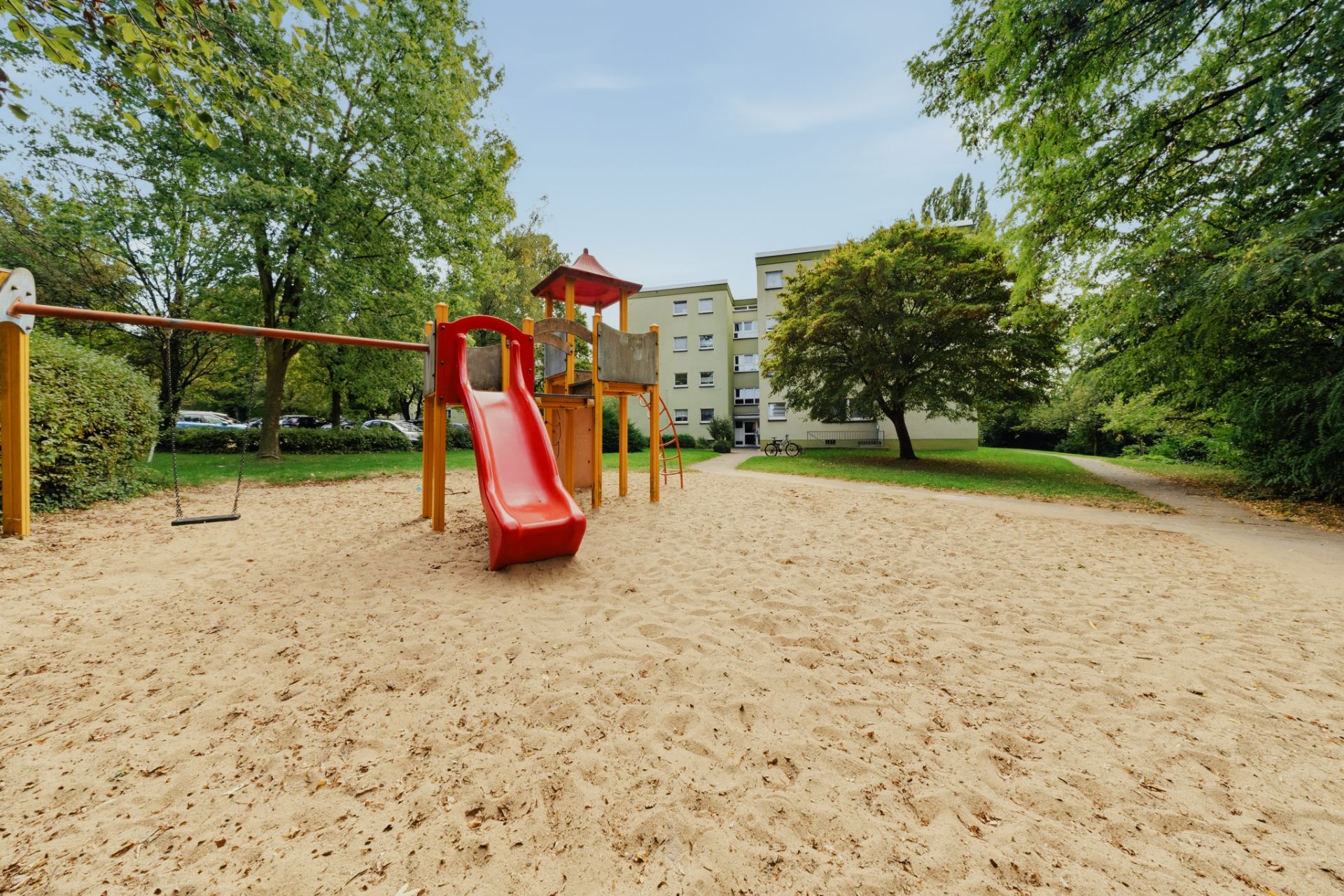 Spielplatz mit Rutsche und Schaukel auf Sand, umgeben von Bäumen und Wohngebäuden.