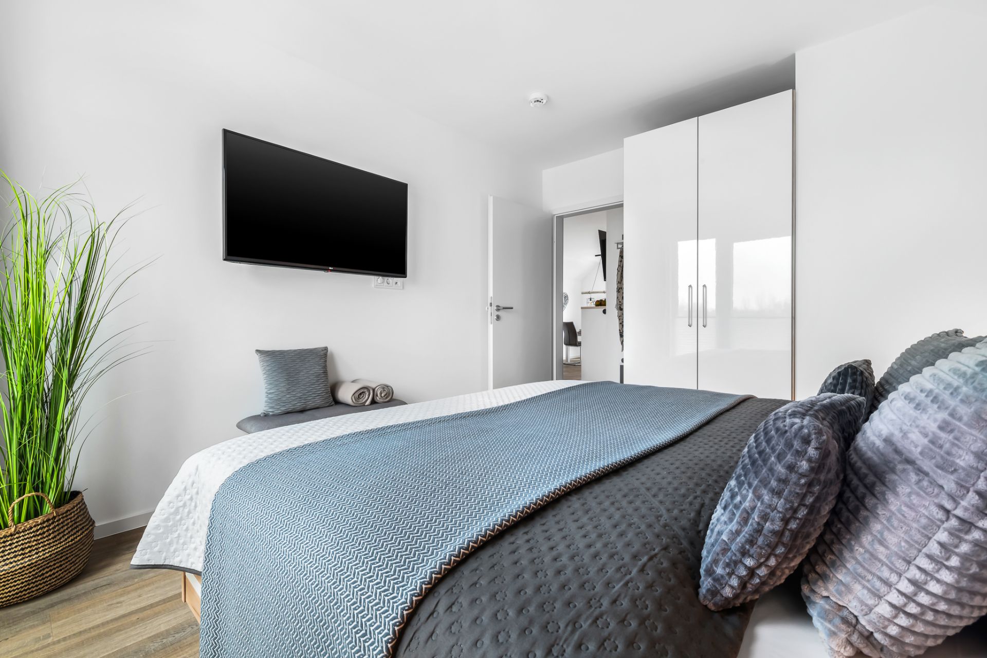 Modernes Schlafzimmer mit Doppelbett, Fernseher an der Wand, Kleiderschrank und großer Zimmerpflanze.