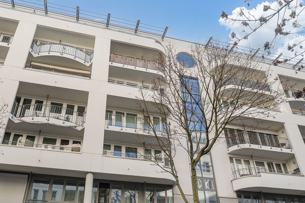 Mehrstöckiges Wohngebäude mit Balkonen und Glasfassade, davor ein kahler Baum.