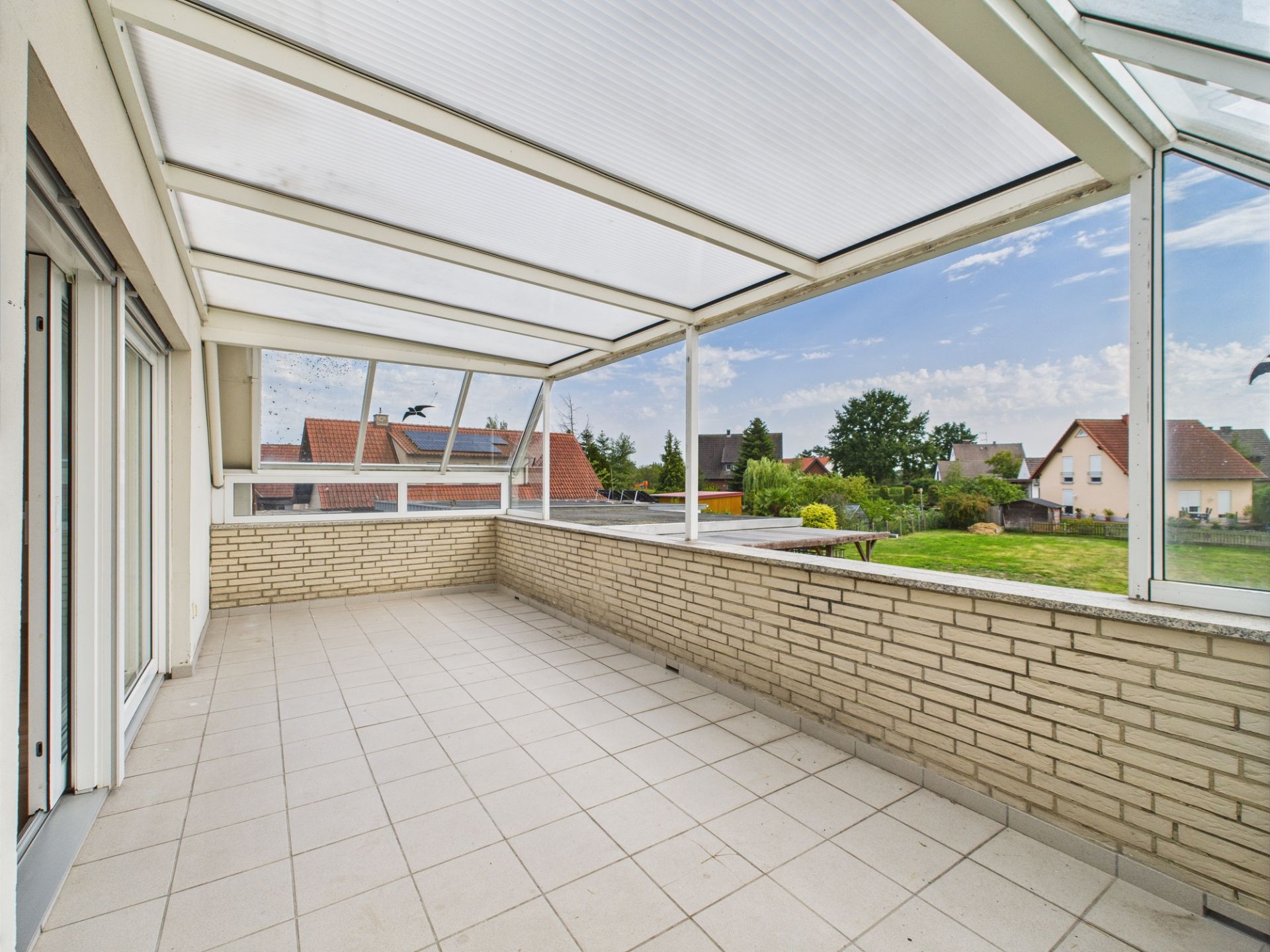 Überdachte Terrasse mit Fliesenboden und Blick auf einen Garten und Nachbarhäuser.