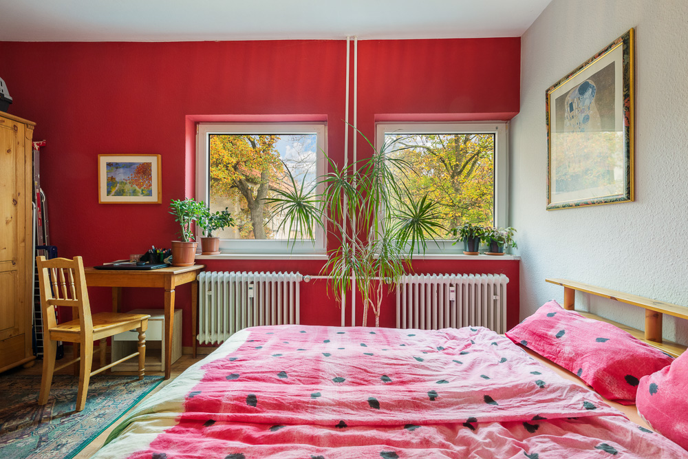 Schlafzimmer mit roter Wand, Doppelbett, Schreibtisch, Holzstuhl und vielen Zimmerpflanzen.