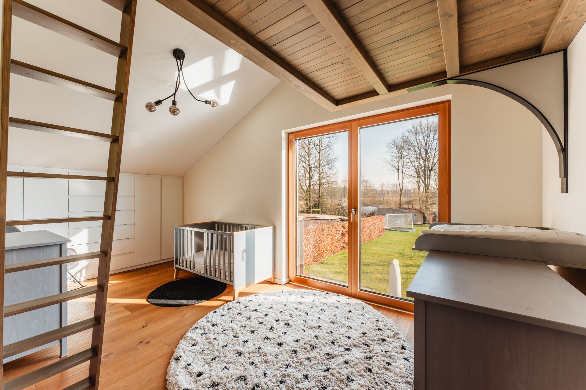 Helles Kinderzimmer mit Holzdecke, Babybett, Wickeltisch und großer Glas-Schiebetür zum Garten.