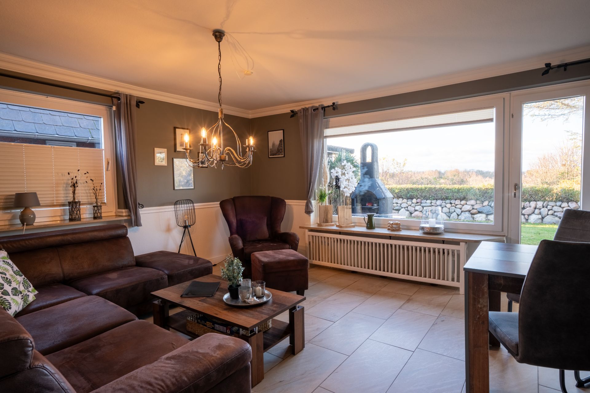 Helles Wohnzimmer mit braunem Sofa, Sessel, Holztisch und großem Fenster mit Blick auf den Garten.