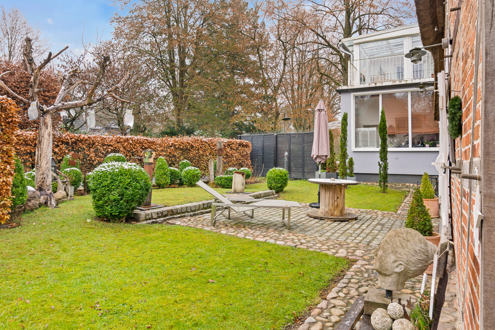 Grüner Garten mit Liegestuhl, runder Tisch, Skulpturen und Büschen neben einem Haus.