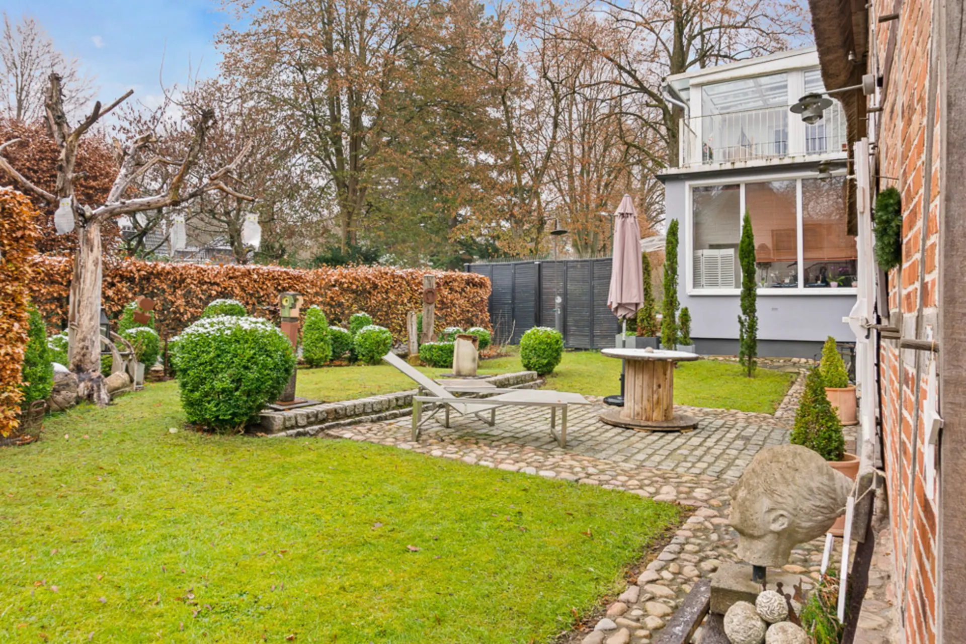 Grüner Garten mit Liegestuhl, runder Tisch, Skulpturen und Büschen neben einem Haus.