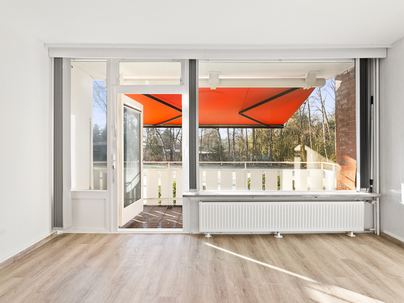 Helles Zimmer mit Holzboden, großer Fensterfront und Zugang zu einem Balkon mit roter Markise.