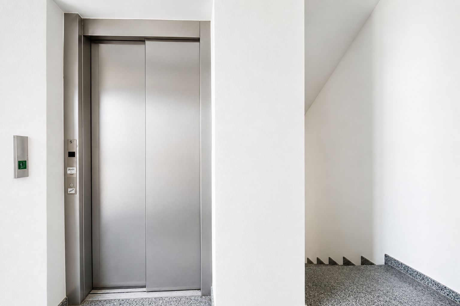 Ein Aufzug mit geschlossener Edelstahltür neben einer Treppe in einem hellen Flur.