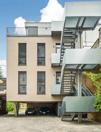 Moderner Wohnblock mit Außentreppe und überdachtem Parkplatz, umgeben von Backsteinwänden und Bäumen.