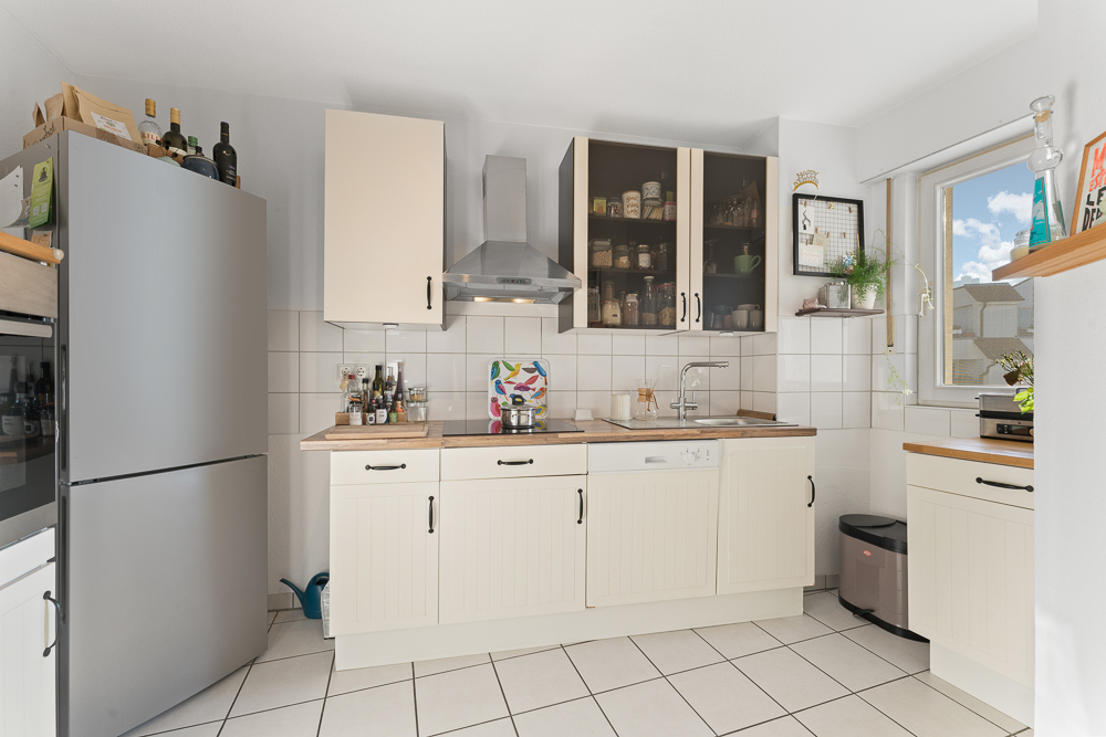 Helle Küche mit weißen Schränken, Kühlschrank, Fenster und offenen Regalen mit Vorratsgläsern.