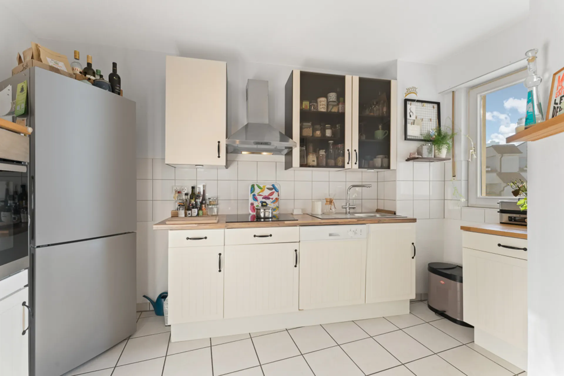 Helle Küche mit weißen Schränken, Kühlschrank, Fenster und offenen Regalen mit Vorratsgläsern.
